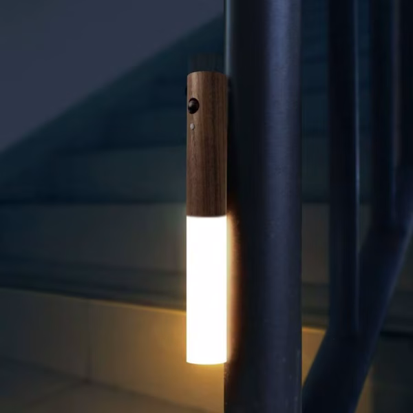 Wooden motion sensor night light mounted vertically on a staircase railing, glowing with warm white light in a dark stairwell