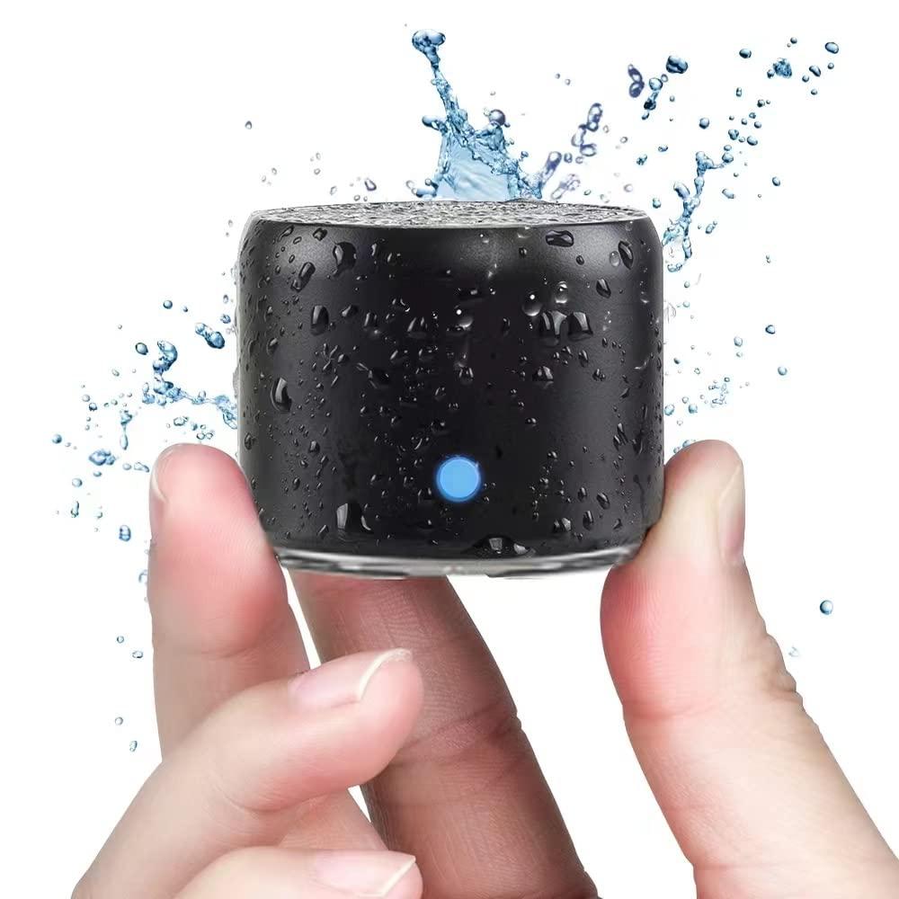A pair of hands holding a small black waterproof Bluetooth speaker with water splashing around it